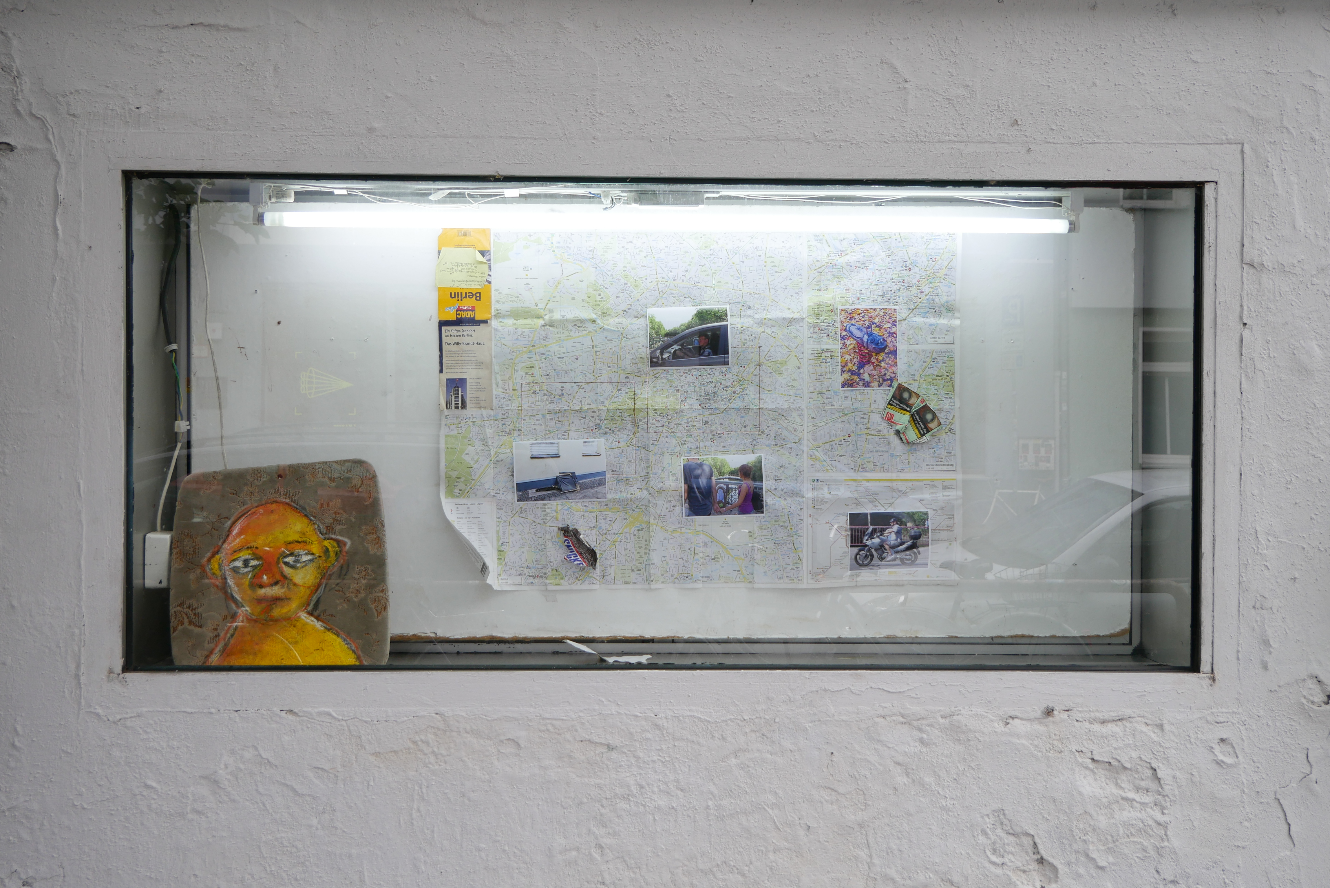 This is an image of a work by the Australian artist Shae Gregg, displayed in the showcase window of the GlogauAIR artist residency in Kreuzberg, Berlin. In it, a map of Berlin is displayed, along with photographs, trash and a painting of a face on an old upholstered chair seat.