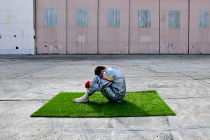 Photograph of a person sat on a square of grass surrounded by an industrial background and concrete ground, by artist Kyle Giacomo, resident at GlogauAIR October 2019.